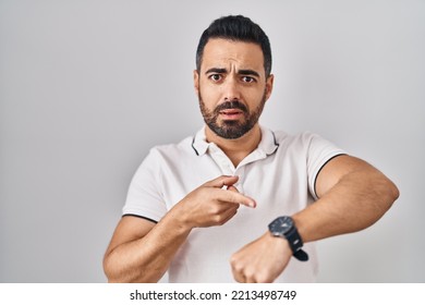 Young hispanic man with beard wearing casual clothes over white background in hurry pointing to watch time, impatience, upset and angry for deadline delay  Foto stock