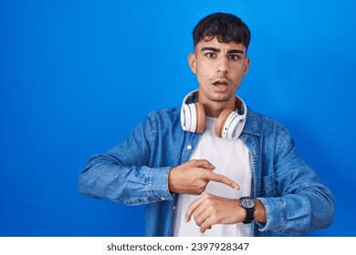 Young hispanic man standing over blue background in hurry pointing to watch time, impatience, upset and angry for deadline delay  Foto stock