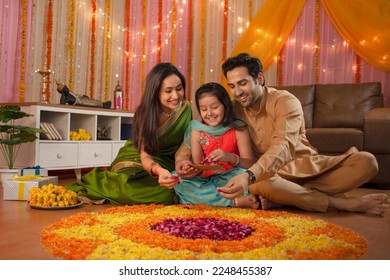 Young Indian family making flower rangoli for Diwali festival celebration. A healthy family sitting together and decorating the house, making flower rangoli, daughter holding flower tray - In... Stock Photo