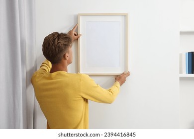 Young man hanging picture frame on white wall indoors, back view: stockfoto