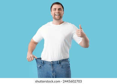 파란색 바탕에 엄지 손가락을 보여주는 느슨한 청바지의 젊은 남자. 체중 감량 개념 스톡 사진