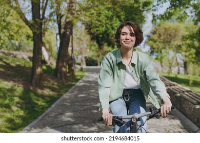 젊은 펜시브 꿈을 꾸는 행복한 여성 20대들이 도시 봄 공원의 인도에서 자전거 자전거를 타는 캐주얼 녹색 재킷 청바지를 입고 옆을 바라본다. 사람들은 도시의 건강한 생활 방식 사이클링 개념을 활발하게 한다 스톡 사진