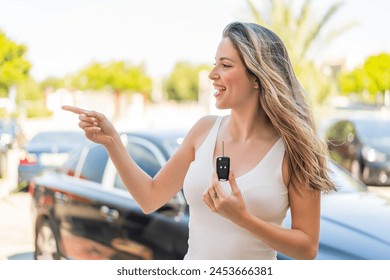 Joven mujer bonita que sostiene las llaves del coche al aire libre que señala al lado para presentar un producto Foto de stock