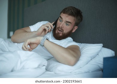 Young sleepy awaken man is just wake up with phone call, looking at his watches checking time and hurry up and yawn. Guy overslept job  Foto stock