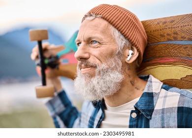 アクティブなクールひげを生やした古いヒップスターの男性はイヤホンを着てスケートボードを持つ自然公園に立っている。自由の精神と極端なスポーツの趣味を楽しんで成熟した旅行者スケーターは、イヤホンで音楽を聴く。の写真素材