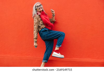 Active senior woman dancing cheerfully against a red background. Happy elderly woman feeling youthful and vibrant while wearing colourful clothing. Excited mature woman celebrating her retirement. 库存照片