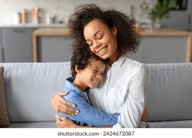 Affectionate black mother hugging her preteen son, both smiling with genuine happiness and warmth, in bright, comfortable living room setting, sitting on sofa ภาพถ่ายสต็อก