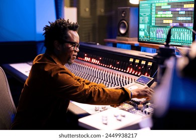 Productor de música afroamericano componiendo Pistas en la sala de control, editando canciones y ajustando Configuraciones de volumen con mandos en el tablero. Ingeniero de audio presionando faders y Botones. Foto de stock