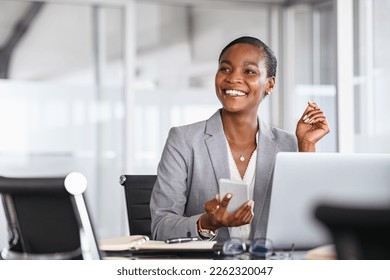 African black business woman using smartphone while working on laptop at office. Smiling mature african american businesswoman looking up while working on phone. Successful woman entrepreneur. Stock-foto