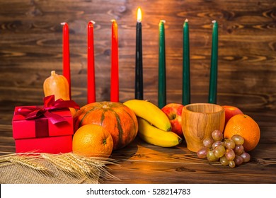 african Kwanzaa festive concept with decorative candles red, black and green, gift box, pumpkins, ears of wheat, grapes, orange, banana, bowl and fruits on wood background: stockfoto
