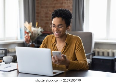 アフリカの女性は、職場のデスクに座ってノートパソコンを見つめて携帯電話を保持し、オフィスでコンピュータとガジェットの間でデータを同期し、企業のデバイスとビジネスアプリケーションを使用し、仕事を計画し、主催者を使用しますの写真素材