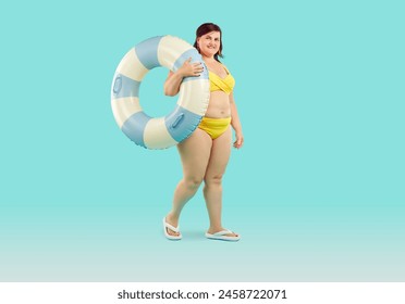 明るい青の背景に水泳サークルでポーズを取った水着で驚くべき肥満ぽっちゃりの女性。スタジオで着色された背景に海で泳ぎに行く太った陽気な女性の全長ポートレートの写真素材