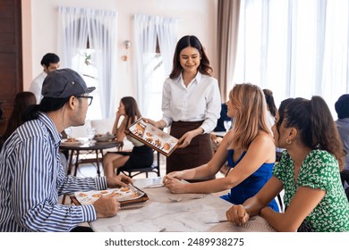 Asian waitress serving food to group of diverse customer in restaurant. Attractive female server service woman work by deliver meal and drink to consumer at their table in dining room with happiness. 库存照片