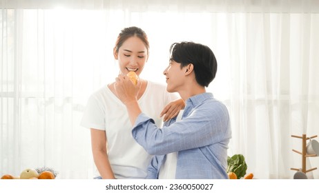 Asian young happy couple boy and girl feeding bread bakery and take care each other in the morning breakfast romantic lifestyle routine with sunrise at the window curtain in a kitchen apartment 库存照片
