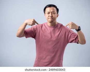 ピンクのTシャツを着たアジア人男性は、明るいグレイのスタジオ背景に立って、自信に満ちた表情で両手を指しています。彼の主張の強い姿勢は自信と誇りを伝えますの写真素材
