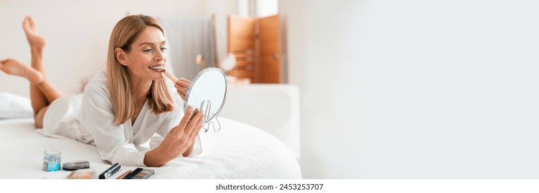 Mujer feliz atractiva de mediana edad que aplica el lápiz labial, que mira el espejo mientras está acostado en la cama y que pone en el maquillaje decorativo, que utiliza el producto cosmético en interiores, panorama con el espacio de la copia Foto de stock