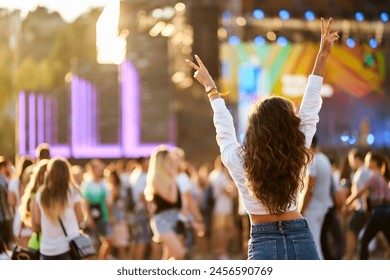 Back view of joyful woman with raised hands enjoys summer music festival. Crowd dances at beach concert, sunset light. Happy fan cheers at outdoor live event. Excited attendee celebrates, vibes. 库存照片