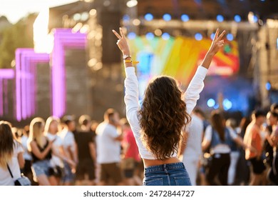 Back view of young woman at summer beach music festival. Raised hands with peace sign, enjoying live concert. Crowd, stage lights, sunset party vibes. Casual fashion, outdoor event, fun atmosphere. 库存照片