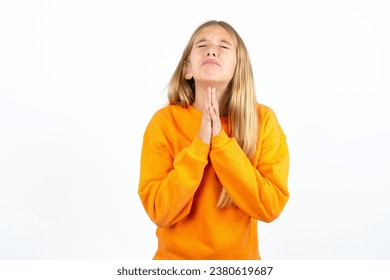 beautiful caucasian kid girl orange sweater using smartphone begging and praying with hands together with hope expression on face very emotional and worried. Please God: stockfoto
