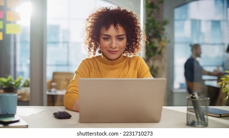 クリエイティブオフィスのデスクに座っている美しい中東部長。マーケティング代理店のラップトップコンピュータを使用して巻き毛の若いスタイリッシュな女性。バックグラウンドで作業する同僚の写真素材