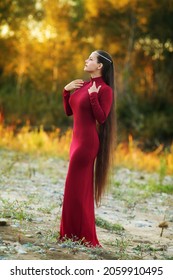 夕暮れの秋の森の背景にぴったりした赤いドレスのポーズを取った、美しいスタイリッシュなブルネット。地面にほとんど触れそうな、華やかでとても長い髪をしたシックな若い女性の写真素材