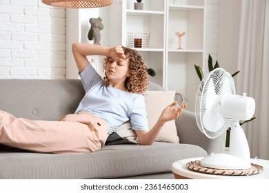 Beautiful young woman with electric fan lying on sofa in living room during heatwave 库存照片