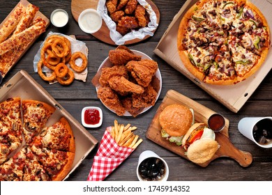 Buffet table scene of take out or delivery foods. Pizza, hamburgers, fried chicken and sides. Above view on a dark wood background. स्टॉक फोटो