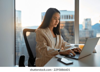 Busy Asian business woman using laptop in company office. Young female digital finance professional worker using computer doing corporate analysis online management sitting at desk, city window view. Arkistovalokuva