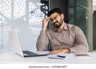 Businessman experiencing stress and headache while working on laptop in a modern office setting, exemplifying work-related strain.: stockfoto