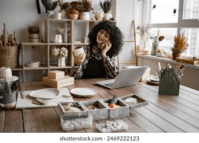 Caucasian female potter entrepreneur talking to buyer of handmade mug sitting at table and taking order using laptop. Successful business lady owner of shop made of clay talking to customer. 库存照片