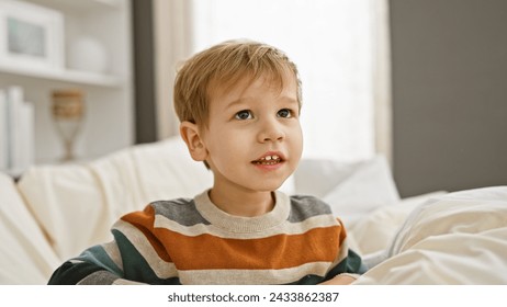 Стоковая фотография: Caucasian toddler boy with blond hair sitting on a white bed in a bright bedroom, looking thoughtful