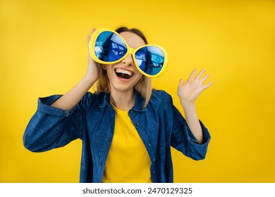 Celebration and party. Having fun. Young pretty woman laughing wearing oversized party glasses wave hello. Colorful studio portrait with yellow background. 库存照片