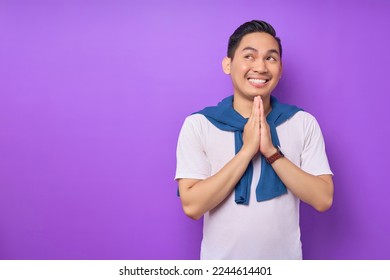 Cheerful young Asian man wearing white t-shirt placing hands in praying gesture and looking aside isolated over purple background. people lifestyle concept: stockfoto