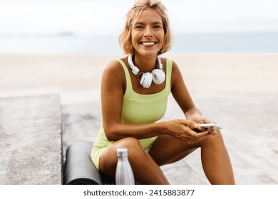 건강하고 활기차고 건강한 생활을 하고 있는 쾌활하고 젊은 백인 여성이 여름날 바다 옆의 계단에 앉아 있다. 해변 요가 운동 후 카메라를 잡고 웃고 있는 여자. 스톡 사진