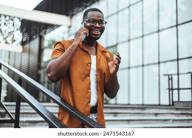 A cheerful black male student talks on his phone while ascending stairs outside a campus building, dressed in casual attire with eyeglasses. Stockfoto