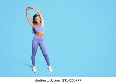 Cheerful millennial caucasian slim lady in sportswear doing arm stretching and warm up exercises isolated on blue studio background, full length. Sports, body care, yoga workout Foto Stock