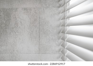 Concept of sunlight and privacy protection on windows in bathroom. Side view of white shutter blinds with wooden slats in tradition venetian style. Copy space on tilled grey wall in loft apartment Foto stock