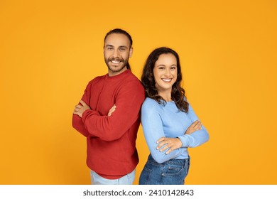 Стоковая фотография: Confident cheerful young man and woman business partners spouses friends standing back to back with arms crossed on chest and smiling at camera, isolated on orange studio background