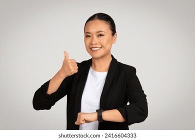 Confiada y contenta empresaria milenaria asiática con el pelo atado, sonriendo y dando un pulgar hacia arriba mientras usa un llamativo y negro, lo que indica aprobación o éxito en un ambiente corporativo. Foto de stock