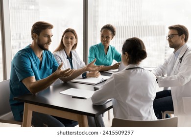 Confident young surgeon man in blue medic uniform talking to multiethnic colleagues in hospital meeting room, speaking to listening diverse doctors at table, offering ideas for clinic teamwork Foto stock