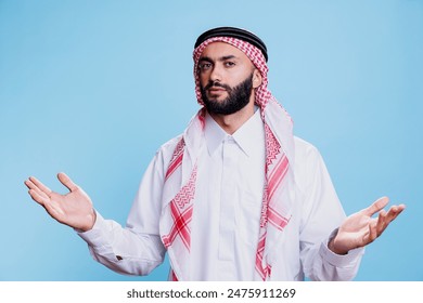 Hombre musulmán confundido en atuendo tradicional extendiendo las manos y expresando incertidumbre retrato. Persona árabe desconcertada encogiéndose de hombros con los brazos abiertos y mirando a la cámara Foto de stock