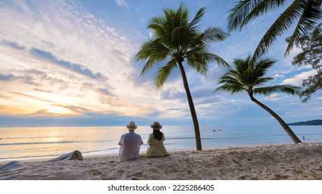A couple of men and women sitting on the beach watching the sunset on the beach with white sand and palm trees, Bang Tao beach Phuket Thailand.  Stock Photo