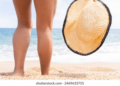 Стоковая фотография: Close up of woman with bare legs holding hat on the shore of a Mediterranean beach on a beautiful summer day