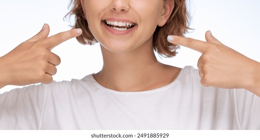 白い背景に笑顔で歯を指さす幸せな女性のクローズアップ画像。歯の健康、前向き、自信を象徴しています。歯科広告、口腔衛生キャンペーンに最適の写真素材