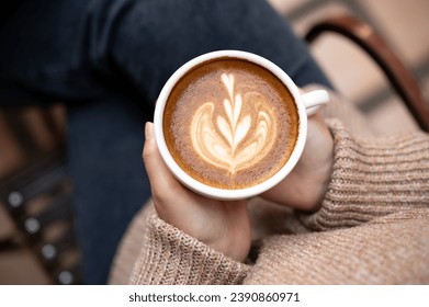 Close-up image of a woman in a cosy knitted sweater holding a cup of hot cappuccino, enjoying morning coffee while sitting at an outdoor table at a cafe in the city in the winter. Foto Stock