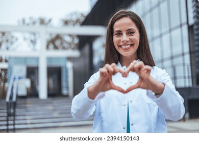Closeup woman nurse making a heart shape with her hands while smiling and standing in hospital. Take care of your heart and love your body. Health and safety in the field of medicine  库存照片
