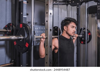 Dedicated and focused young male athlete doing back squats on a smith machine in a well-equipped gym, showing strength and concentration. Stock Photo
