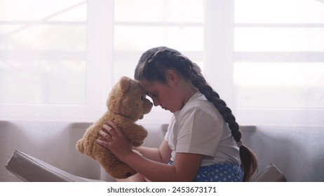 彼女のお気に入りのおもちゃのテディベアと窓のそばに座って落ち込んでいる女の子が動揺します。家族の概念におけるドメスティックバイオレンスの問題。落ち込んでいる女の子は悲しく、クマと窓に日光を退屈していますの写真素材