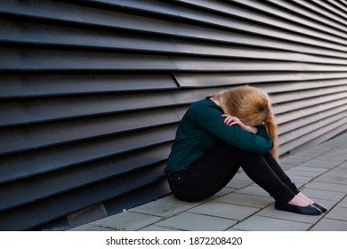 頭を腕に当てた黒い裂け目のある壁に寄りかかる、都会の歩道に座っている落ち込んだ若い女性の写真素材
