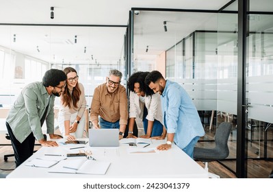 Diverse team huddles around table, and looks at a laptop screen in a bright office, closely reviewing documents and digital content together, showcasing a moment of collective focus and determination Foto stock